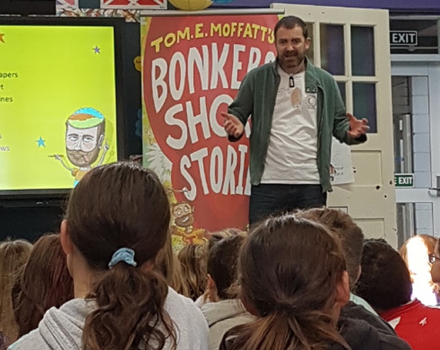 A man in a green jacket stands on stage, facing an audience, with a colorful banner that reads "Bonkers Short Stories.