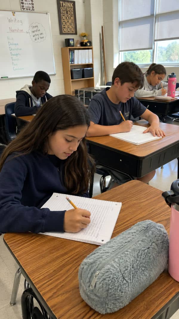 Students working on writing assignments at desks in a classroom, with a gray pencil case and a pink water bottle visible.